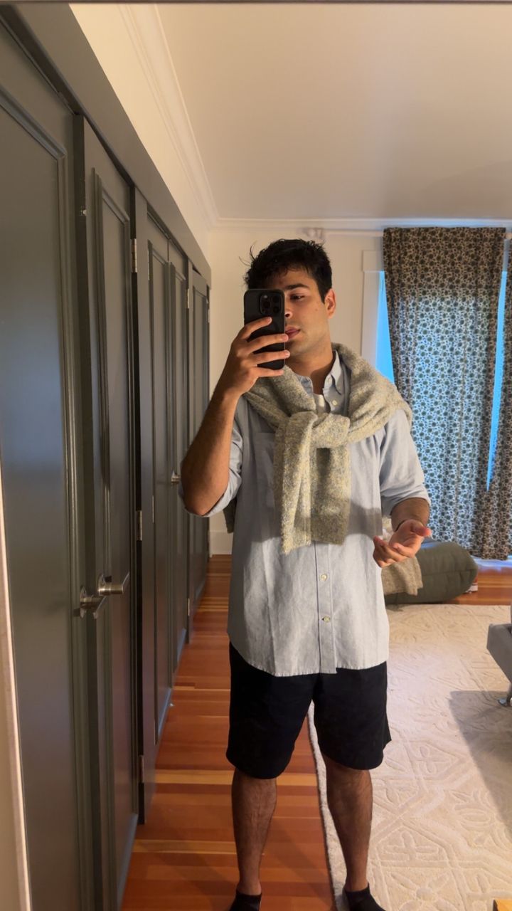 Janav in a mirror selfie wearing the outfit EDIT picked: light blue oxford shirt, grey knit sweater over the shoulders, navy shorts
