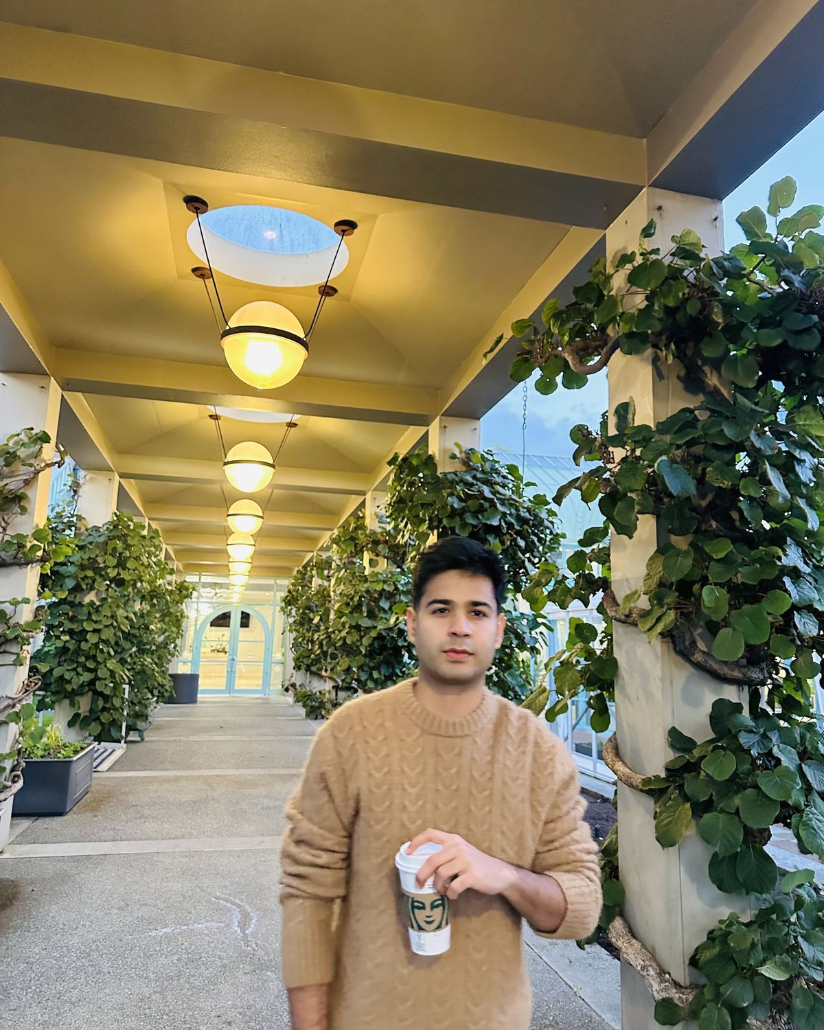 Janav in a tan sweater holding a coffee, in a long plant-lined walkway